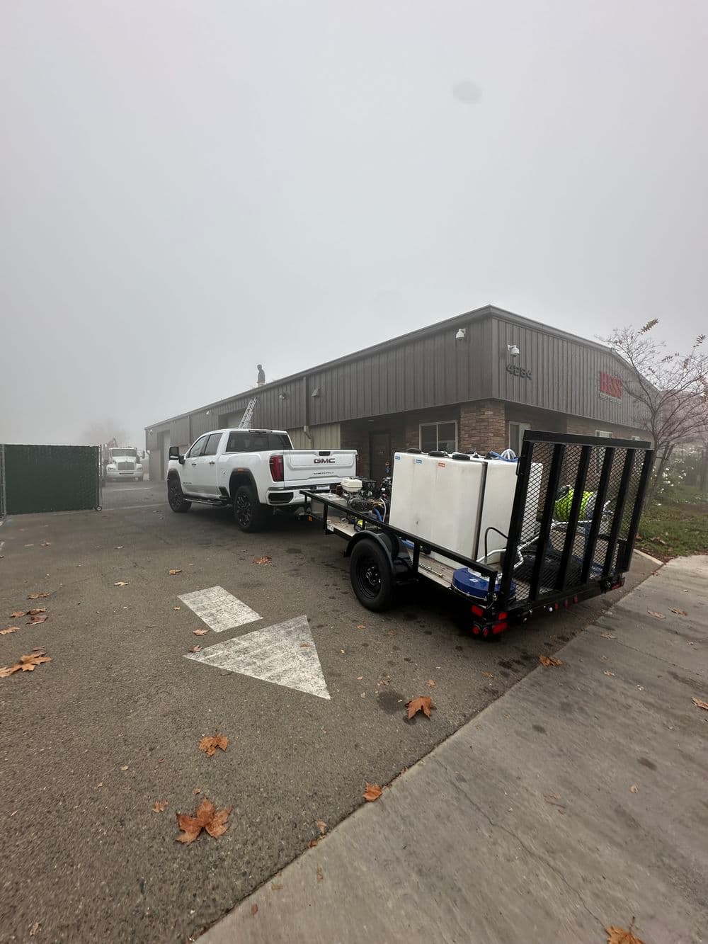 White pickup truck towing a trailer with equipment outside a misty building.
