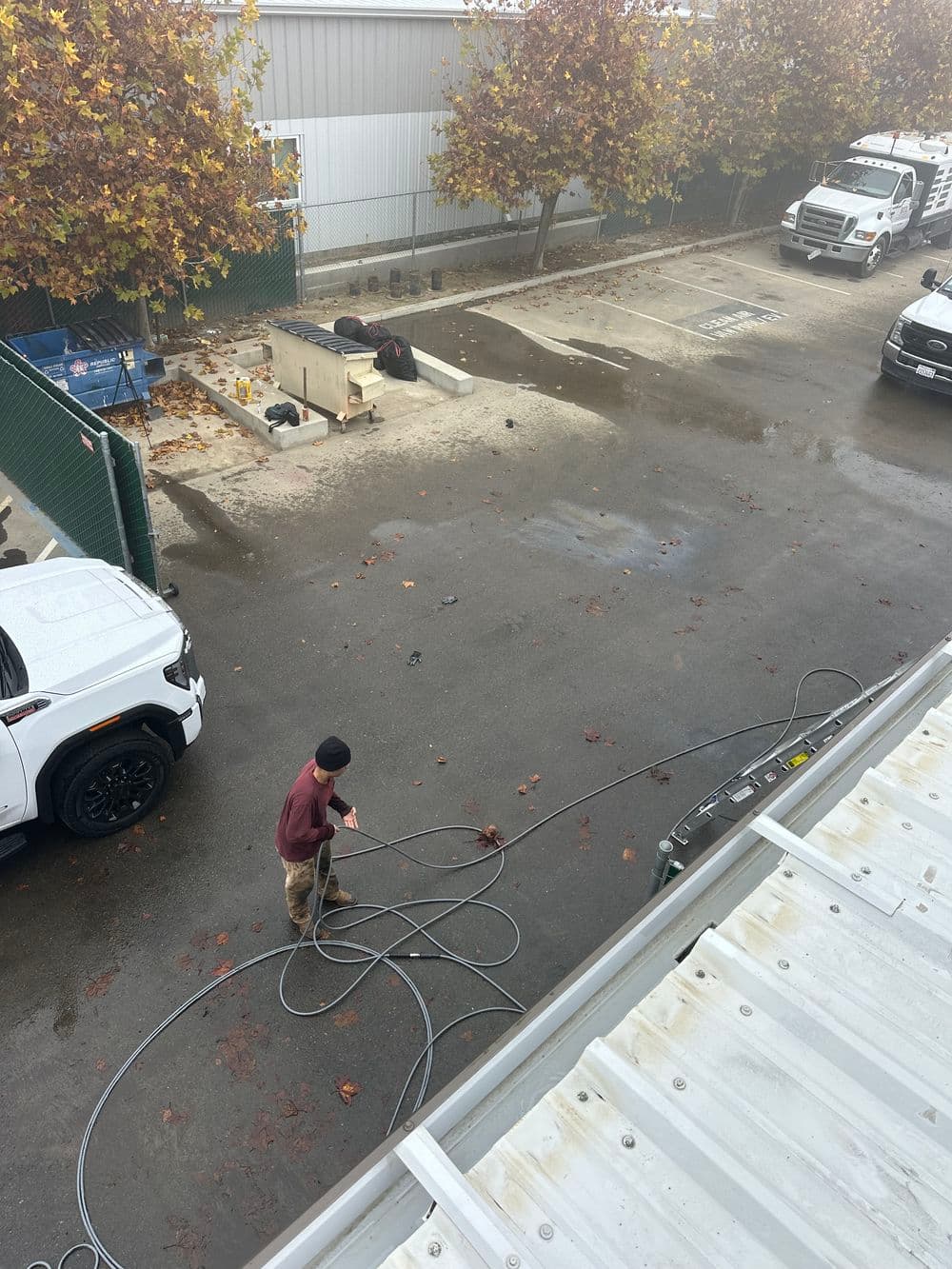 Worker using a power tool in a parking lot with fallen leaves and vehicles nearby.